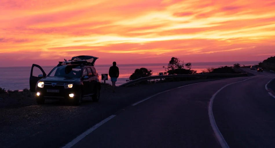 Surfer au Maroc : SUV avec planches de surf sur la côte au coucher du soleil, un surfeur contemple l'océan.