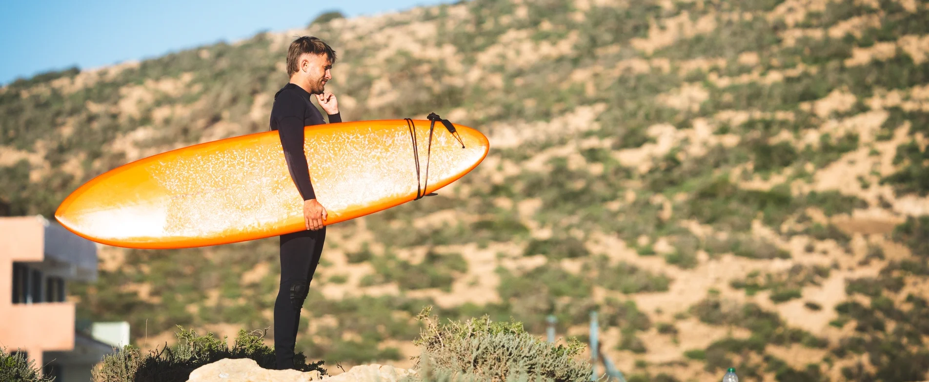 Surfeur en combinaison avec planche orange, prêt à surfer au Maroc. Évaluer le coût d'un surf camp au Maroc.